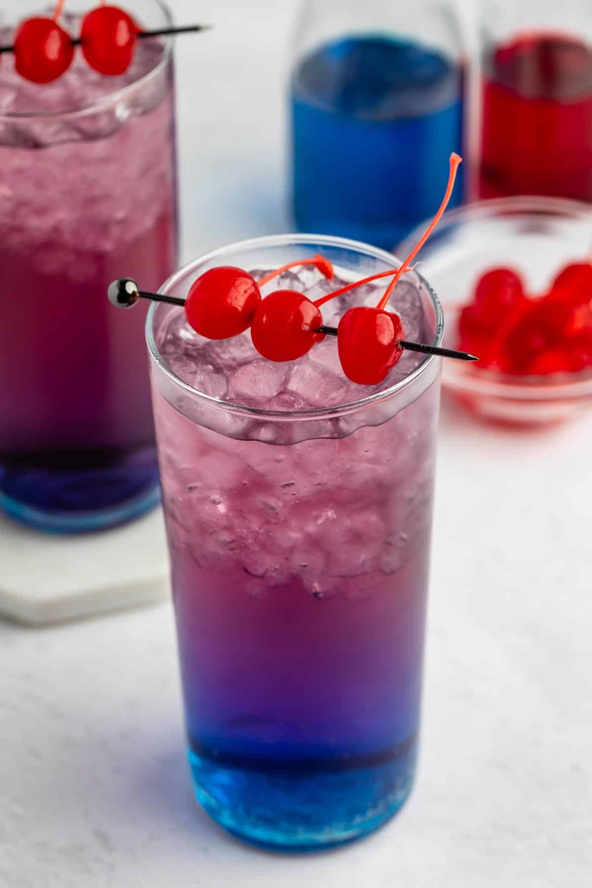 A tall glass filled with blue and purple layered iced drink, garnished with three maraschino cherries on a black stick. In the background, another similar drink and a small bowl of cherries are visible.