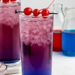 A tall glass filled with crushed ice and a layered blue and purple drink, garnished with three red cherries on a skewer. Bottles of blue and red liquid are in the background.