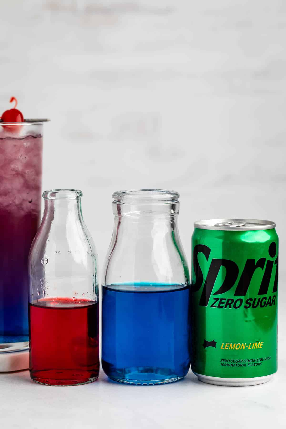 A can of Sprite Zero Sugar sits next to two small glass bottles containing red and blue liquid, with a tall glass of purple drink topped with a cherry in the background.