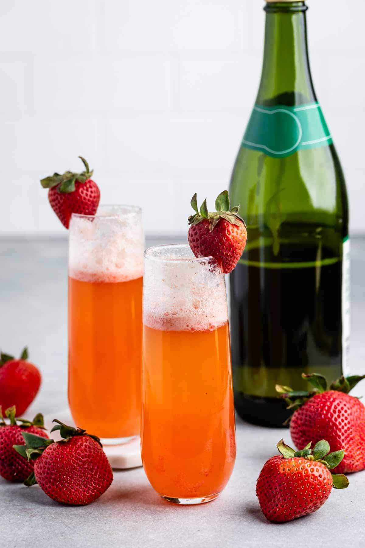 Two glasses of orange-red sparkling drink with foam on top, each garnished with a fresh strawberry, sit beside a green champagne bottle and several scattered strawberries on a light surface.