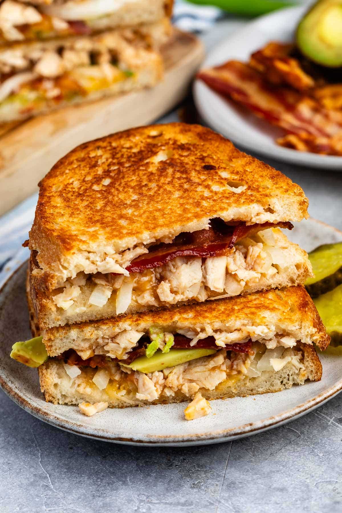 A plate with a stacked grilled sandwich filled with chicken, bacon, onions, and pickles. Slices of pickle are on the side, and more sandwiches and bacon are in the background.