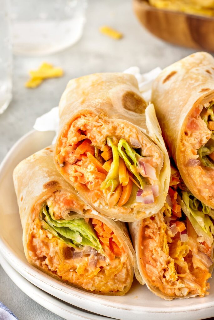 Three tortilla wraps filled with shredded chicken, lettuce, carrots, and red onion, stacked on a white plate. The wraps have a creamy orange filling and are cut in half to show the colorful ingredients inside.