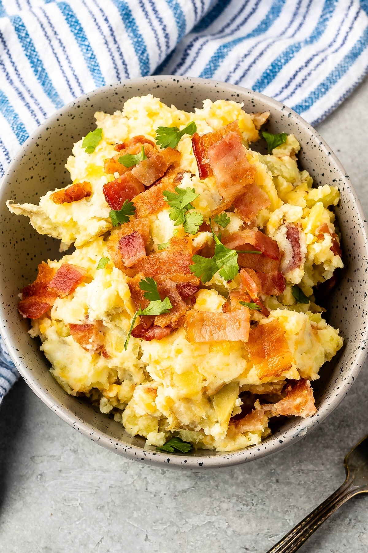 A bowl of mashed potatoes topped with crispy bacon pieces and chopped parsley, placed on a light gray surface with a blue and white striped cloth in the background.