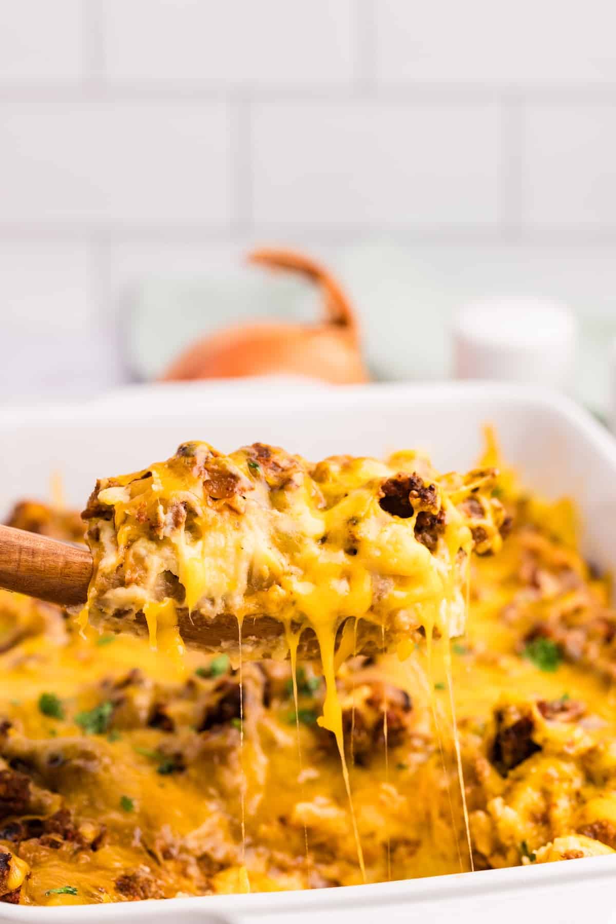 A wooden spoon lifts a cheesy casserole with melted cheese stretching, over a white baking dish filled with the same casserole. An onion and spices are blurred in the background.