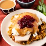 A plate with slices of turkey, mashed potatoes, gravy, and cranberry sauce on stuffing, with a cup of gravy and a bowl of cranberry sauce in the background on a rust-colored tablecloth.