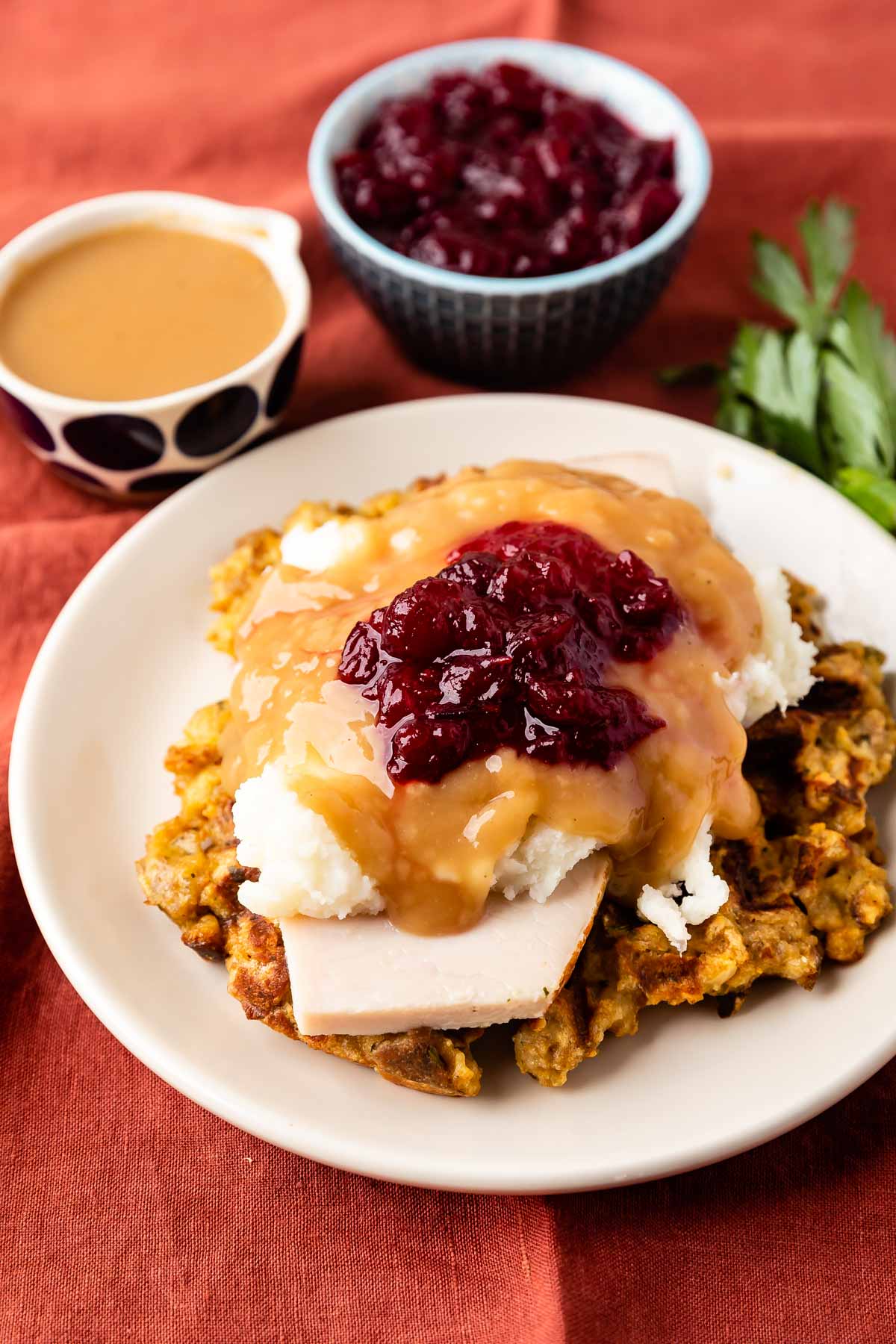 A plate with slices of turkey, mashed potatoes, gravy, and cranberry sauce on stuffing, with a cup of gravy and a bowl of cranberry sauce in the background on a rust-colored tablecloth.