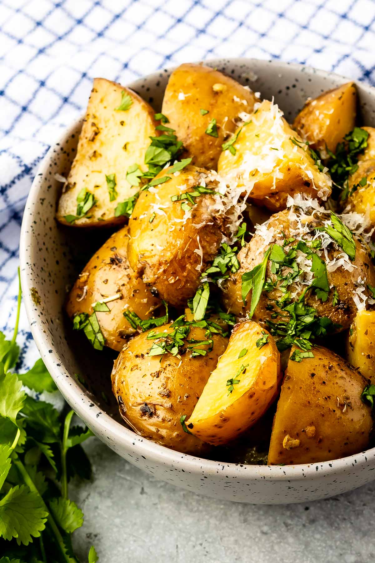 A bowl of roasted potato wedges topped with grated cheese and chopped fresh herbs, placed on a table next to a blue and white checkered cloth and fresh cilantro leaves.