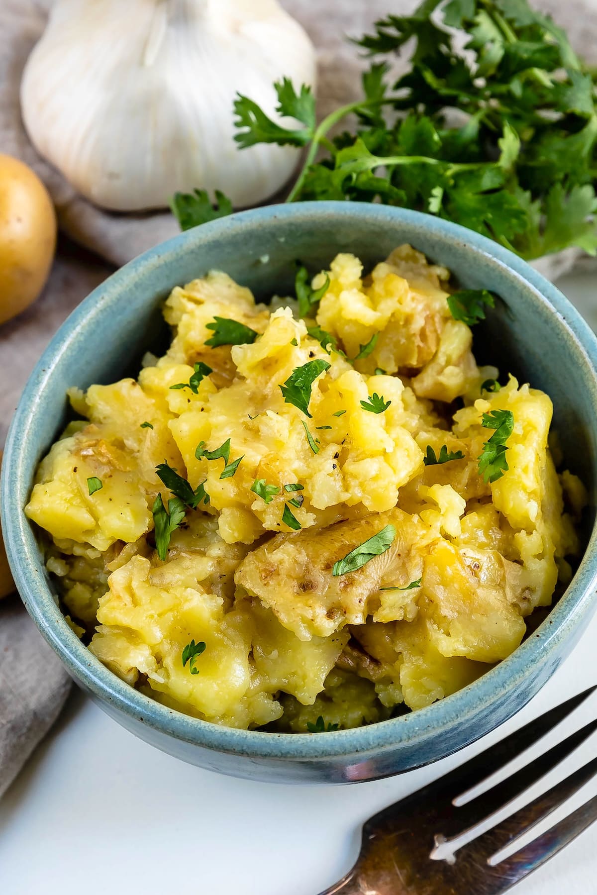 A bowl of mashed potatoes garnished with chopped parsley sits beside a fork, fresh garlic, potatoes, and parsley on a white surface.