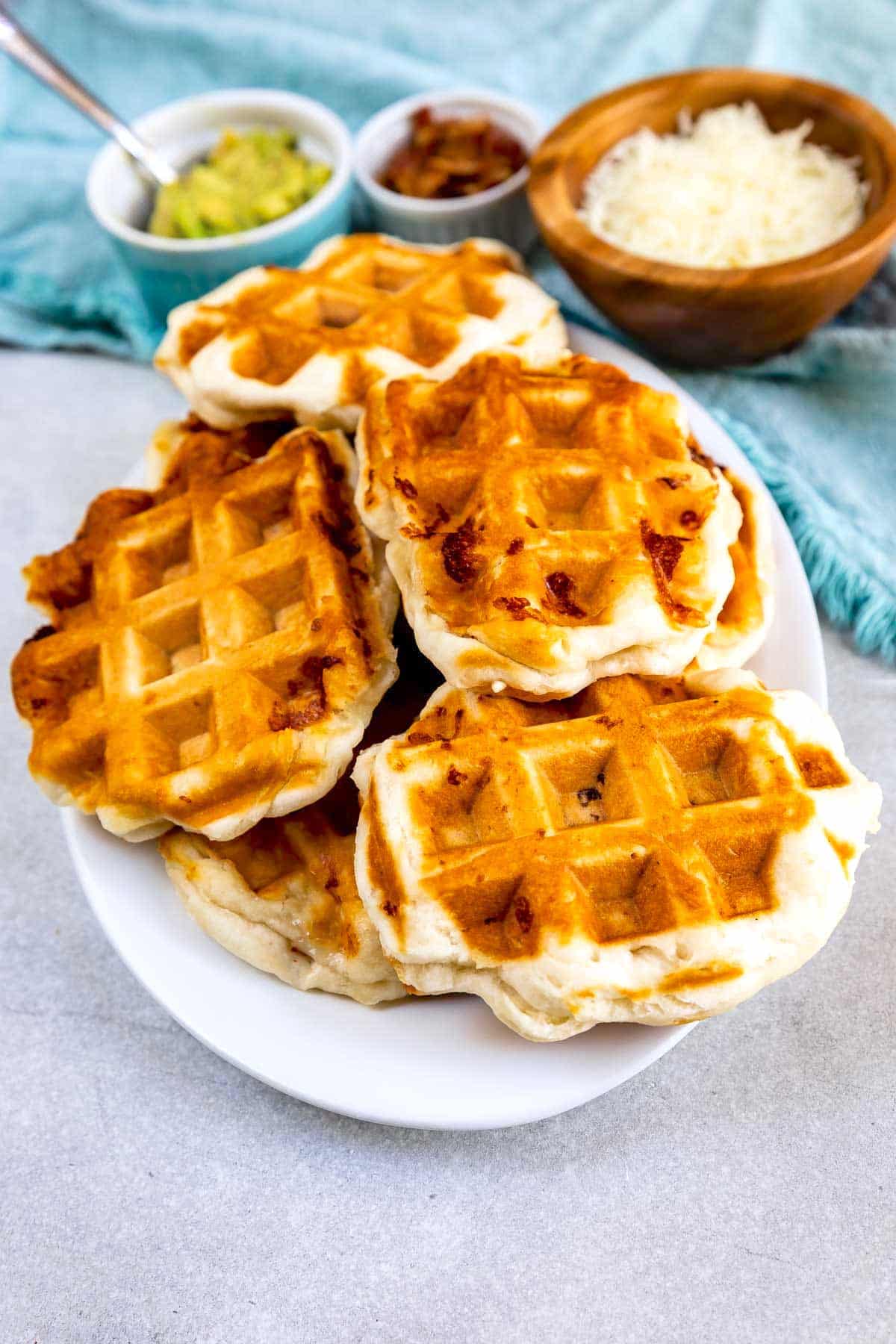 A white plate stacked with golden-brown waffles sits on a light surface. In the background, small bowls contain grated cheese, guacamole, and bacon bits. A blue cloth is partially visible.