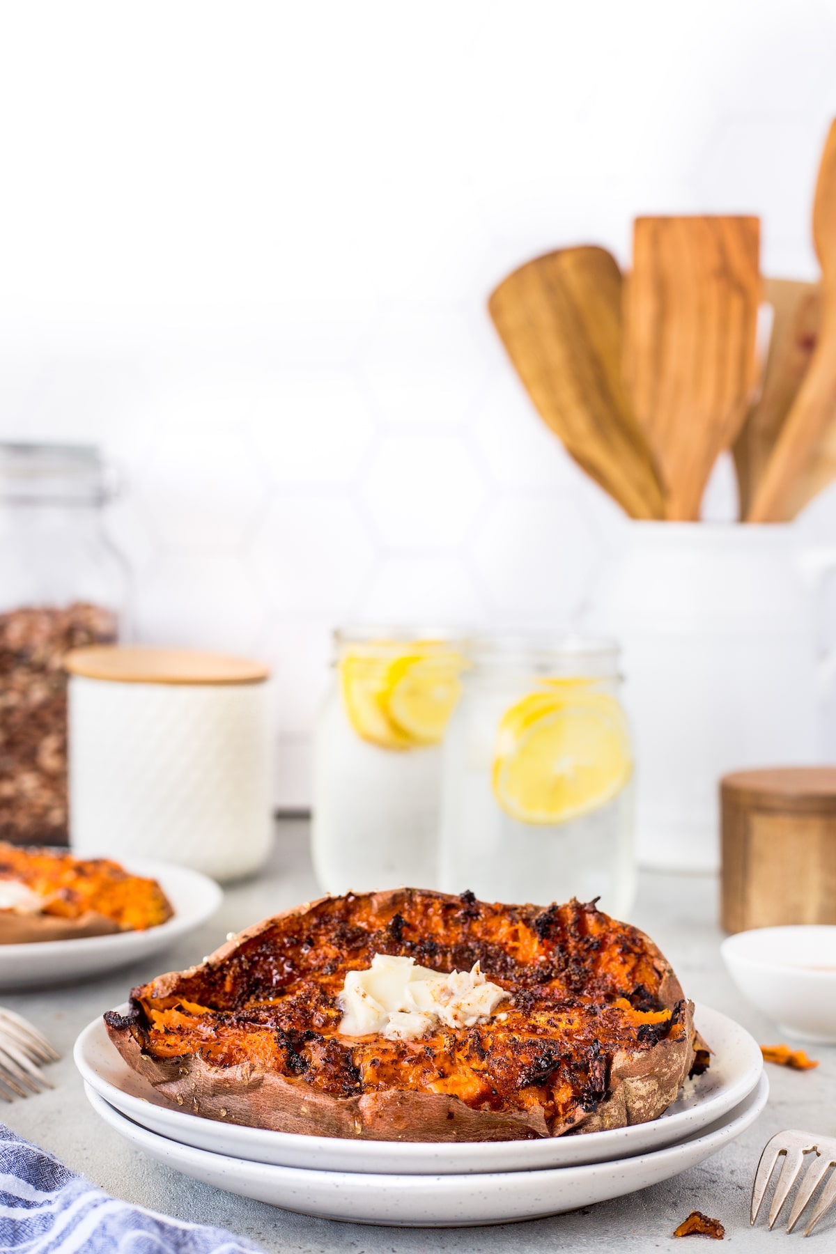 A baked sweet potato topped with butter sits on a white plate. In the background, two mason jars with lemon water, wooden utensils in a holder, and kitchen containers are visible on a light countertop.