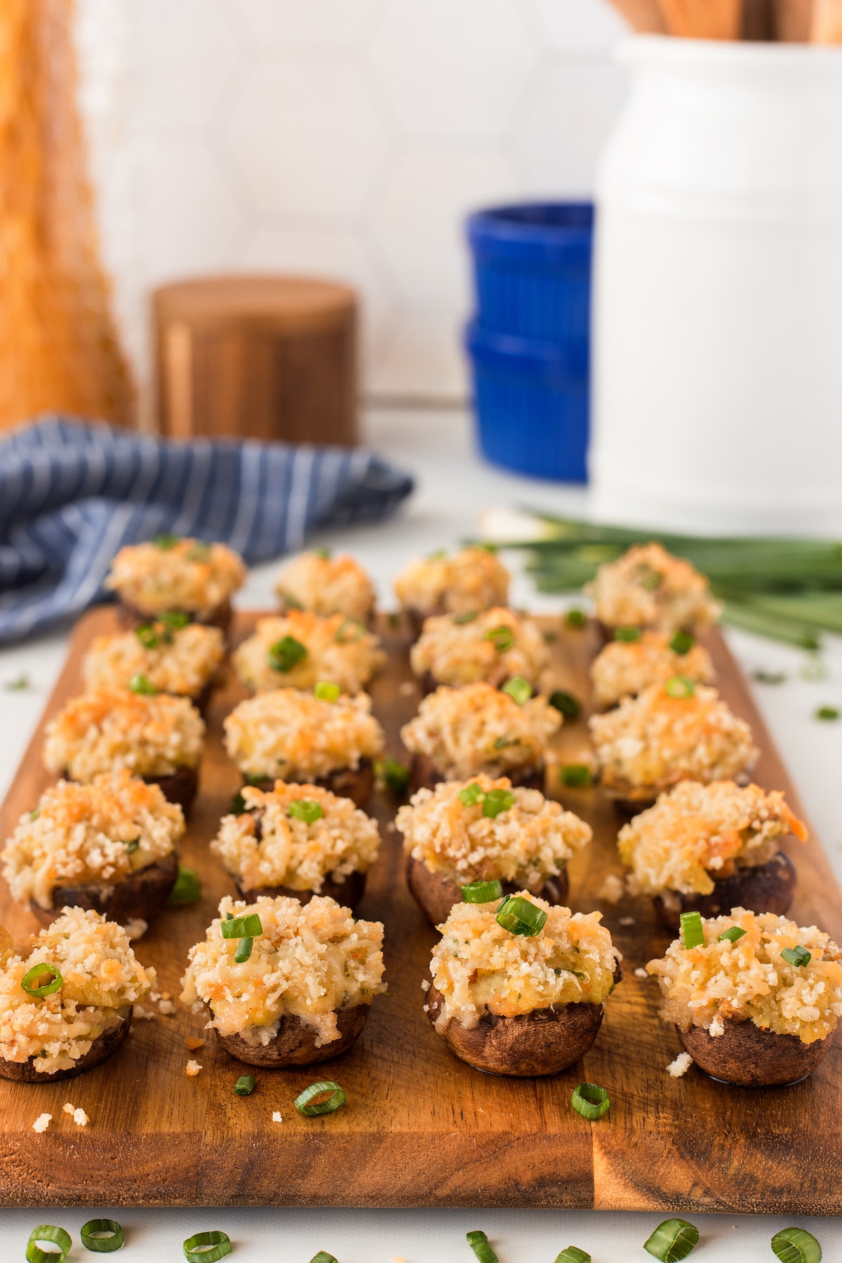 A wooden tray holds several stuffed mushrooms topped with golden breadcrumbs and chopped green onions, arranged neatly with a kitchen setting in the background.