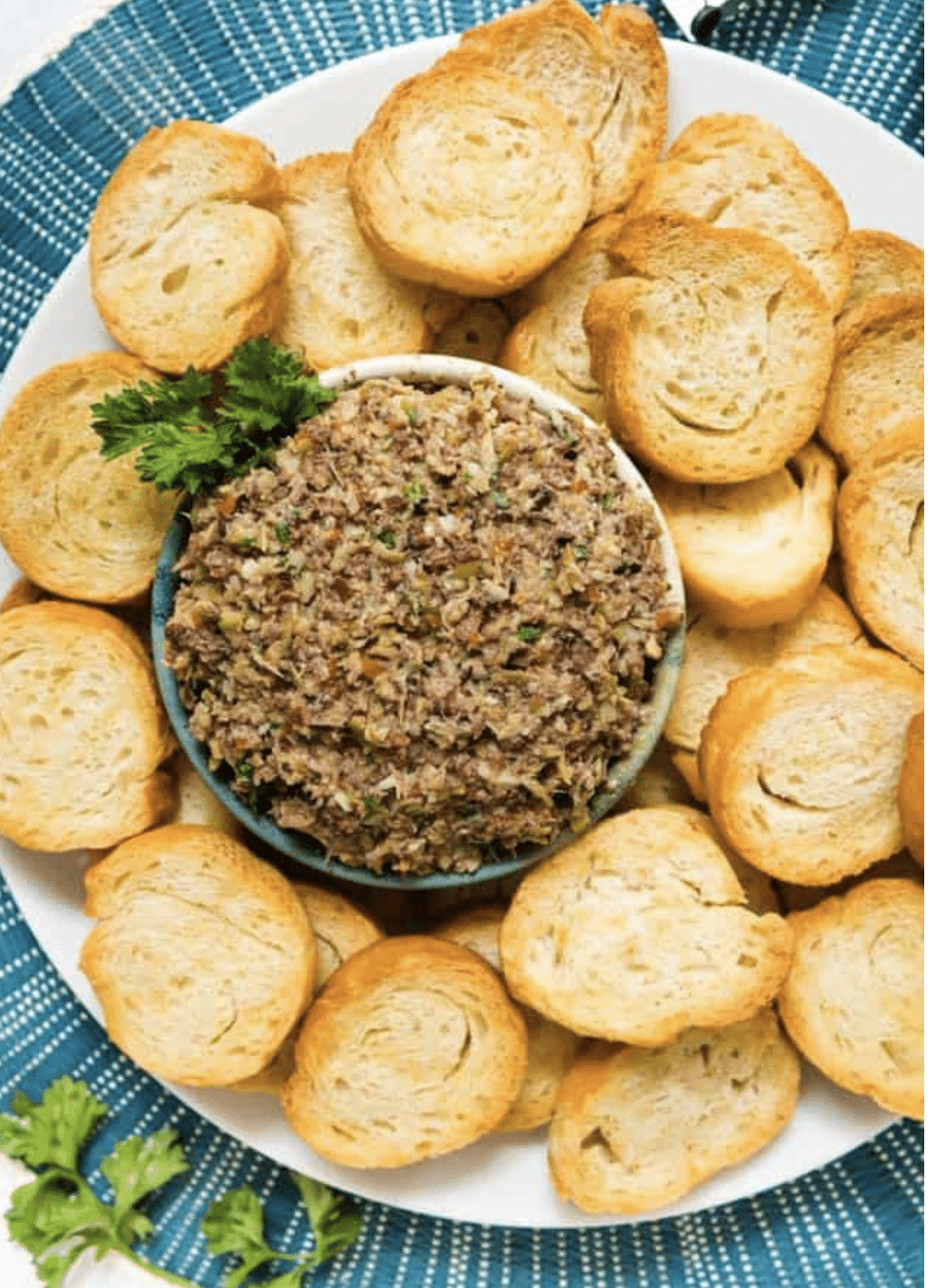 A plate of toasted baguette slices surrounds a bowl of olive tapenade, garnished with parsley, on a blue patterned placemat.