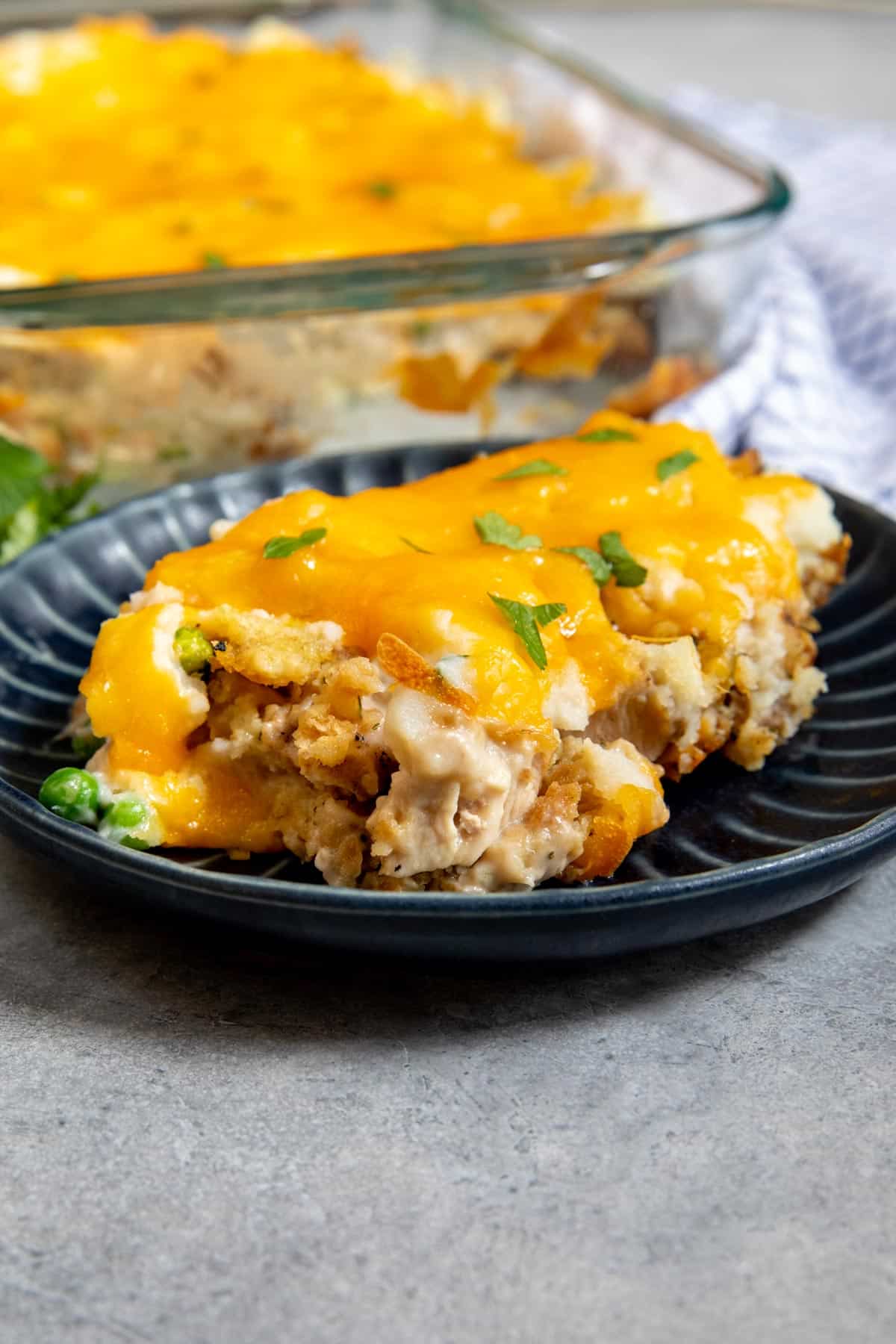 A serving of cheesy chicken casserole topped with melted cheddar and garnished with parsley is on a dark plate, with the rest of the casserole in a glass baking dish in the background.