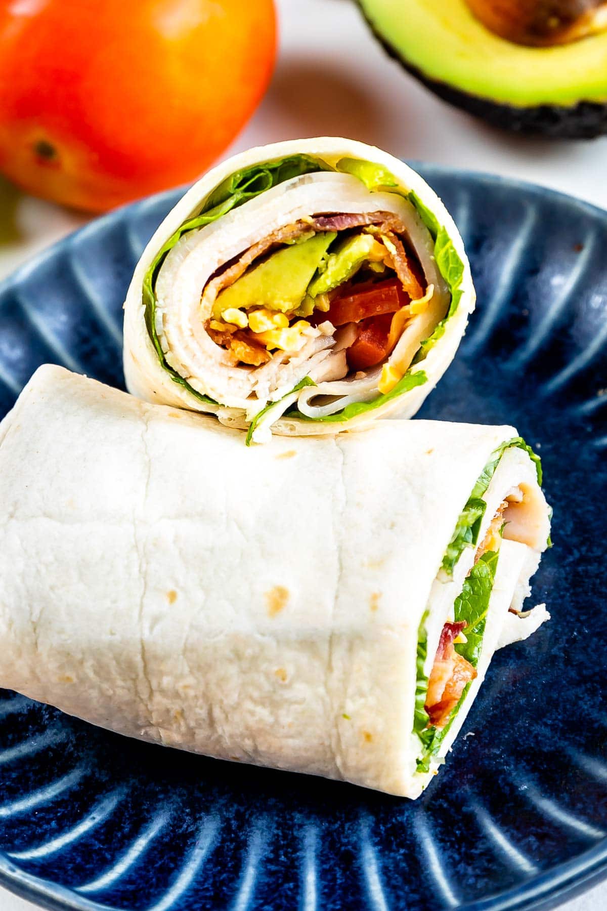 A turkey and veggie wrap cut in half, showing layers of turkey, avocado, lettuce, and tomato, placed on a dark blue plate. In the background, there is a tomato and half an avocado.