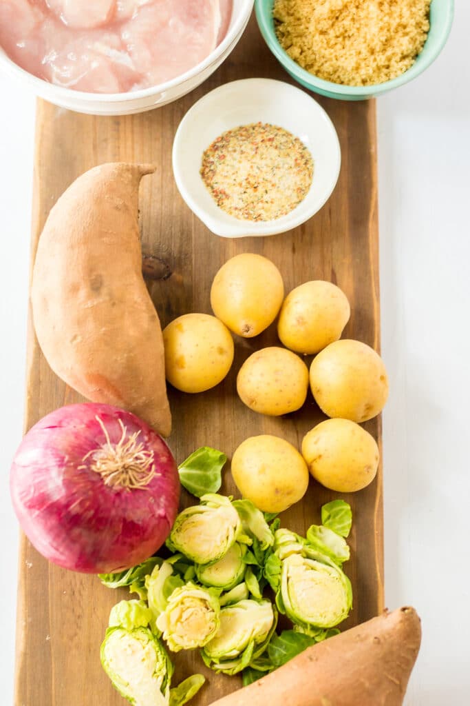 A wooden board with raw sweet potatoes, yellow potatoes, Brussels sprouts, a red onion, and bowls of chicken pieces, breadcrumbs, and seasoning.