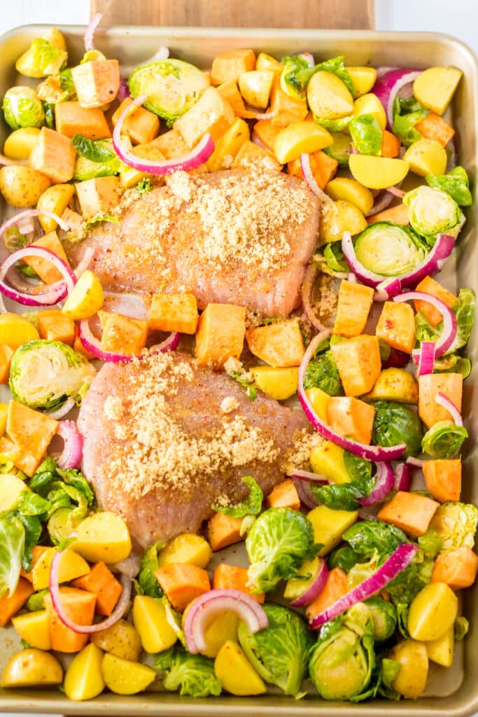 A sheet pan with two raw, breaded chicken breasts surrounded by chopped Brussels sprouts, sweet potatoes, yellow potatoes, and red onion slices, all ready to be roasted.
