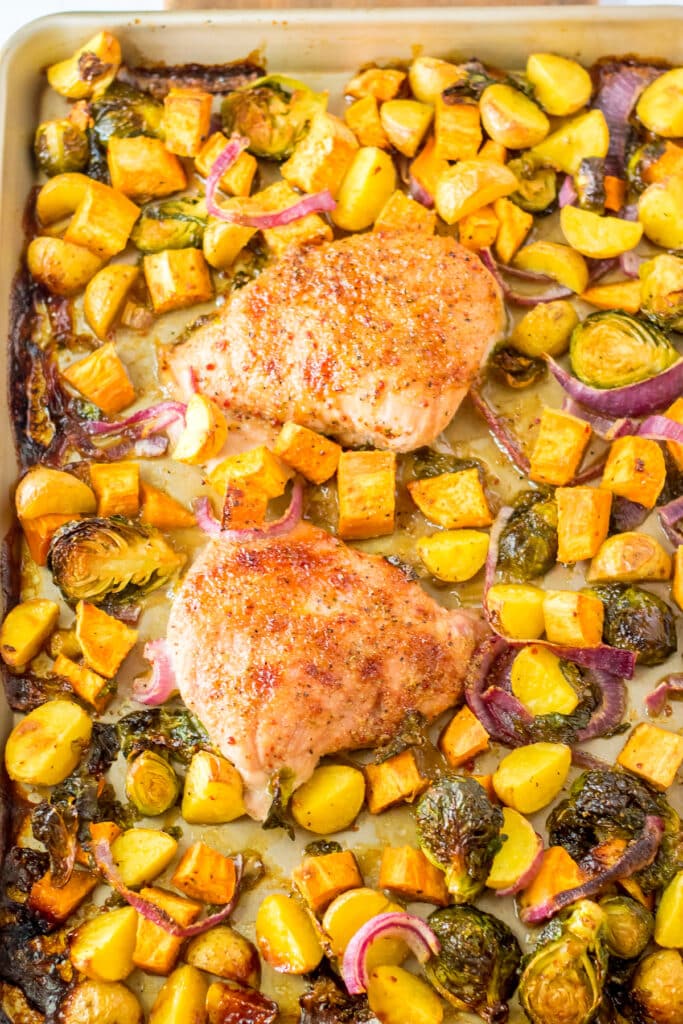 Sheet pan with two seasoned chicken breasts surrounded by roasted yellow potatoes, Brussels sprouts, red onion slices, and chunks of sweet potato. The ingredients are golden and slightly crispy.