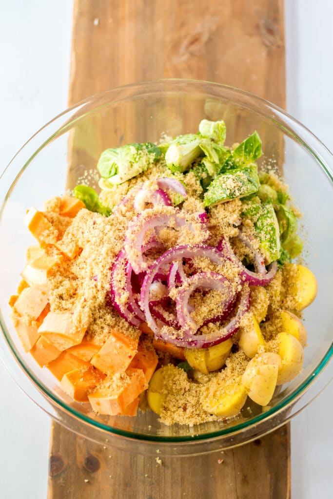 A glass bowl on a wooden surface filled with chopped sweet potatoes, Brussels sprouts, yellow potatoes, red onion slices, and sprinkled breadcrumbs.