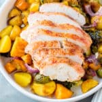 Sliced roasted chicken breast served on a white plate with roasted potatoes, Brussels sprouts, red onions, and carrots on a gray surface.