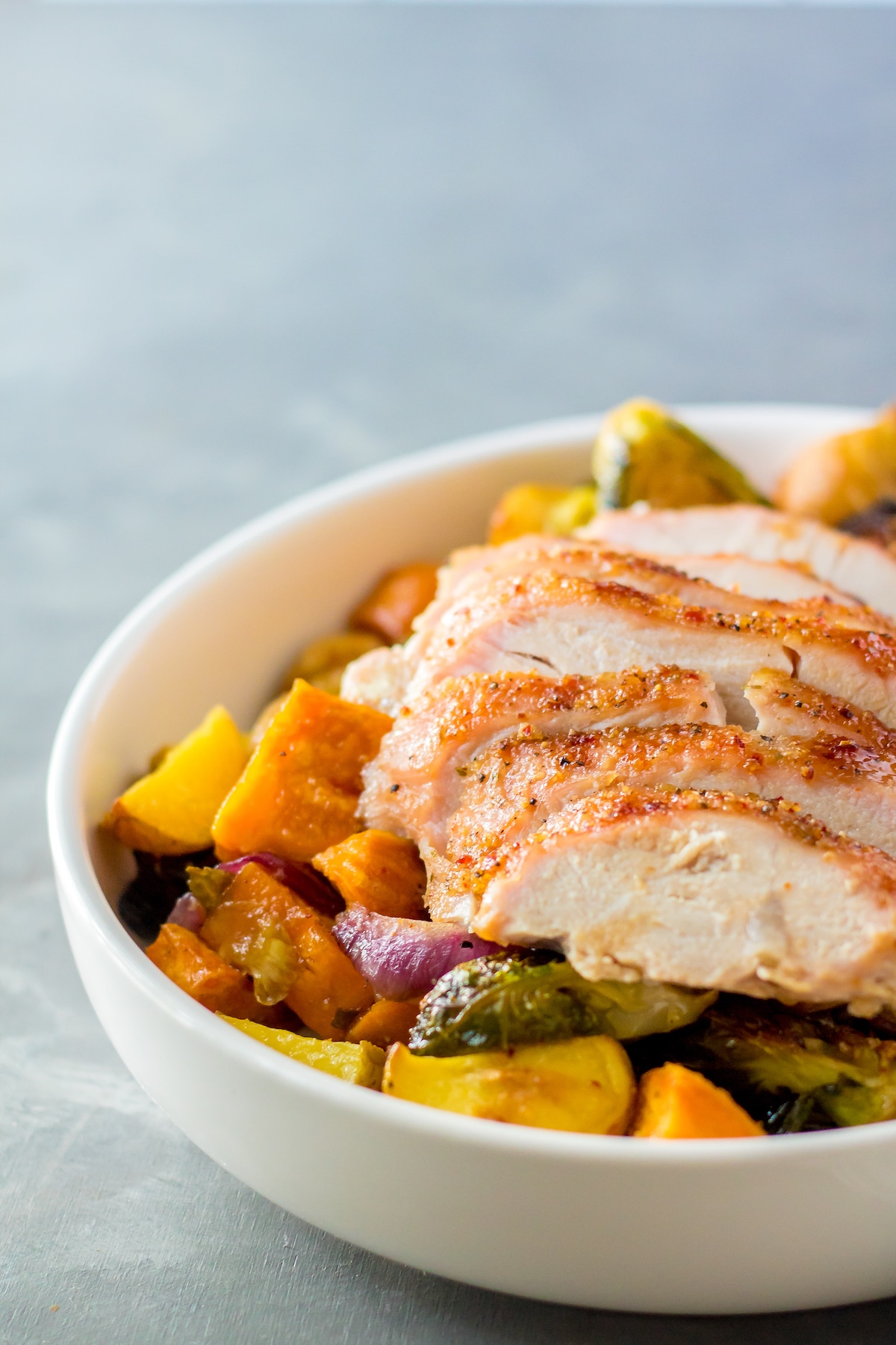 A white bowl filled with sliced roasted chicken breast served over roasted vegetables, including potatoes, brussels sprouts, and red onions. The food appears fresh and appetizing on a light gray background.