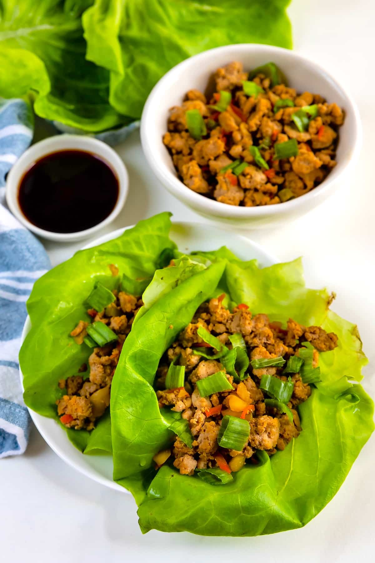 Two lettuce leaves filled with seasoned ground meat and chopped vegetables on a white plate, with extra filling in a bowl and a small dish of dark dipping sauce nearby.