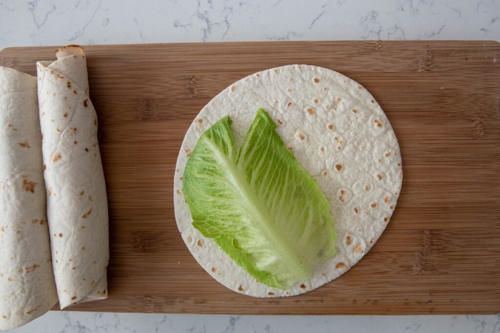 A large lettuce leaf is placed on a flour tortilla on a wooden cutting board, with two rolled-up tortillas nearby. The scene is set on a white marble surface.