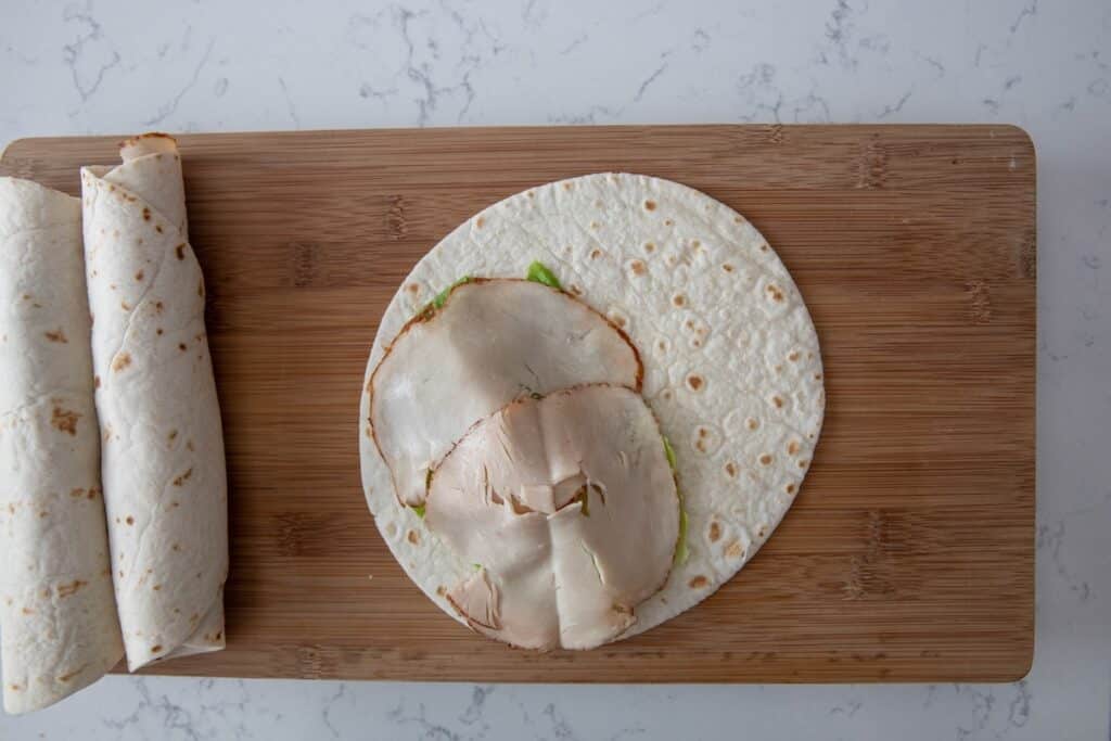 A tortilla with slices of deli turkey and lettuce is open on a wooden cutting board, next to two rolled-up wraps. The background is a white marble surface.