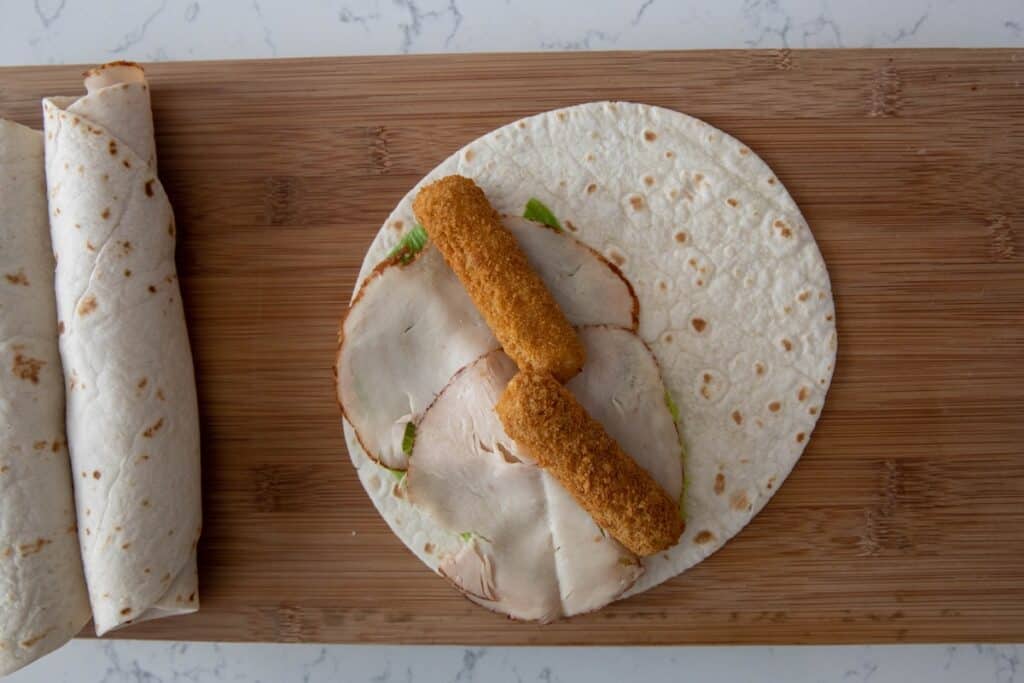 A tortilla wrap on a wooden board topped with slices of deli turkey, lettuce, and two breaded fish sticks. A completed wrap is rolled up and placed to the left.