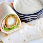 A close-up of a tortilla wrap filled with lettuce and a breaded, fried item, served on a white plate next to a bowl of creamy dipping sauce.