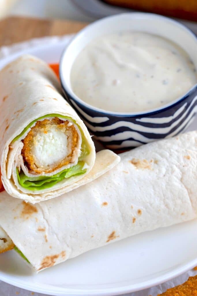 A close-up of a tortilla wrap filled with lettuce and a breaded, fried item, served on a white plate next to a bowl of creamy dipping sauce.