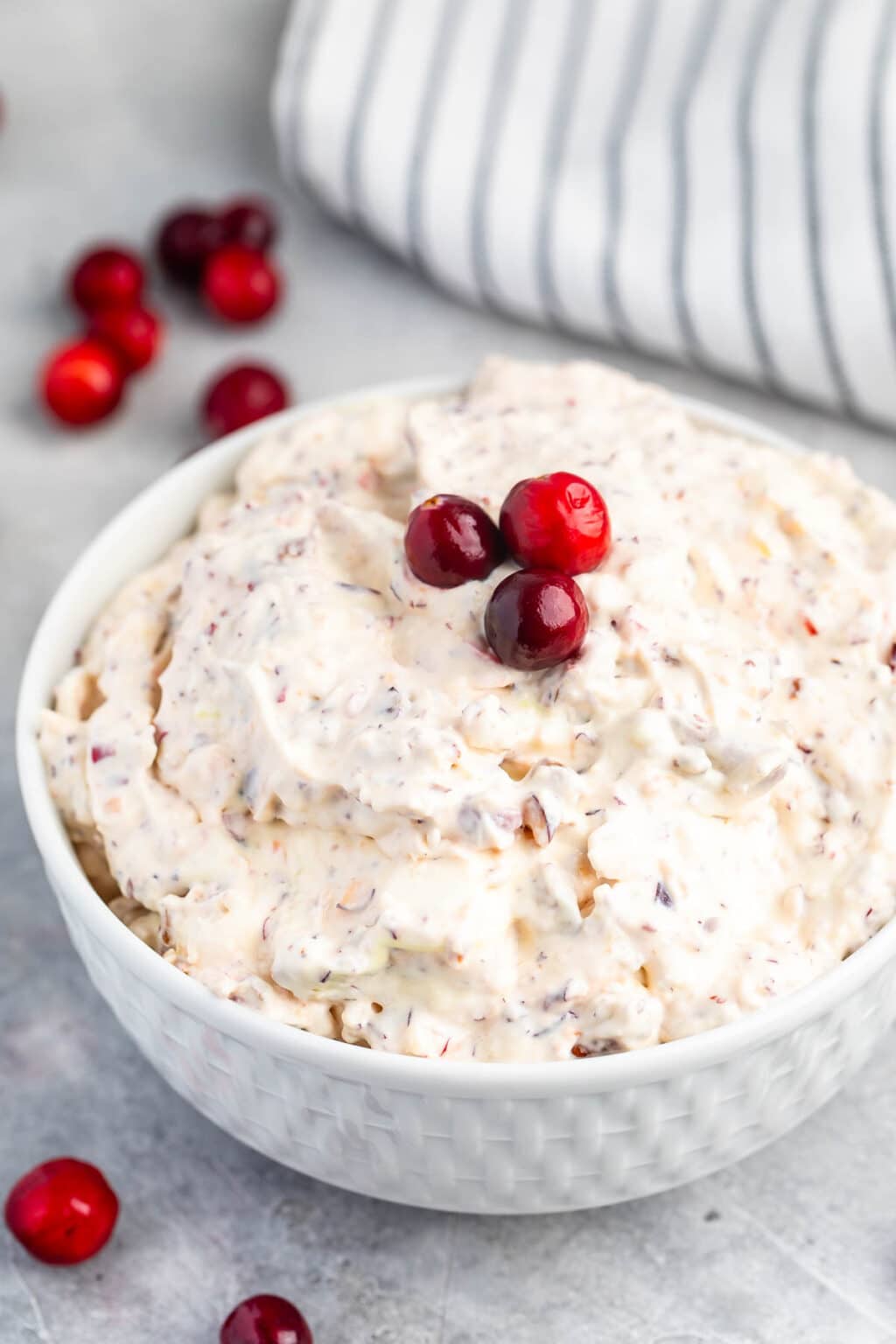 A white bowl filled with creamy cranberry fluff salad, topped with three whole cranberries, sits on a light surface with loose cranberries and a striped cloth in the background.