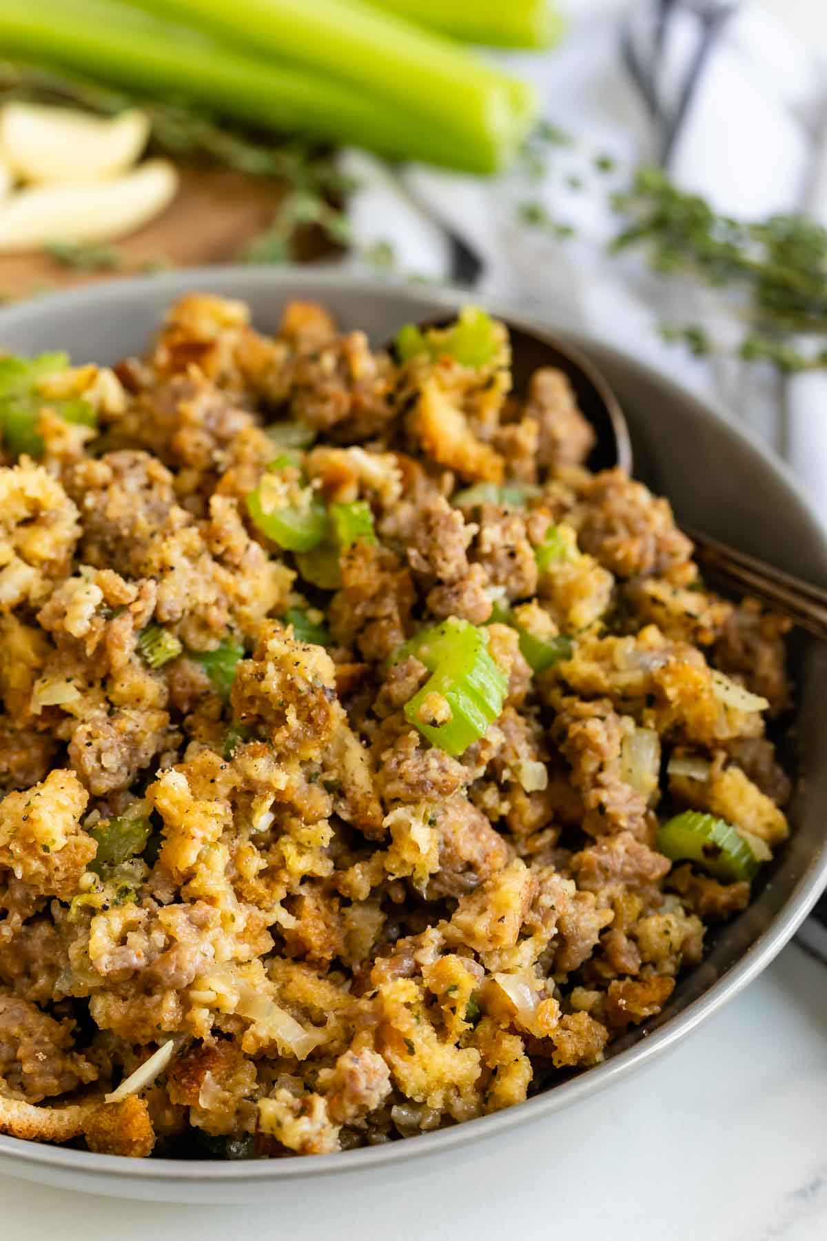 A bowl filled with homemade stuffing containing crumbled sausage, celery, onions, and bread cubes, garnished with herbs, with fresh celery stalks and herbs in the background.
