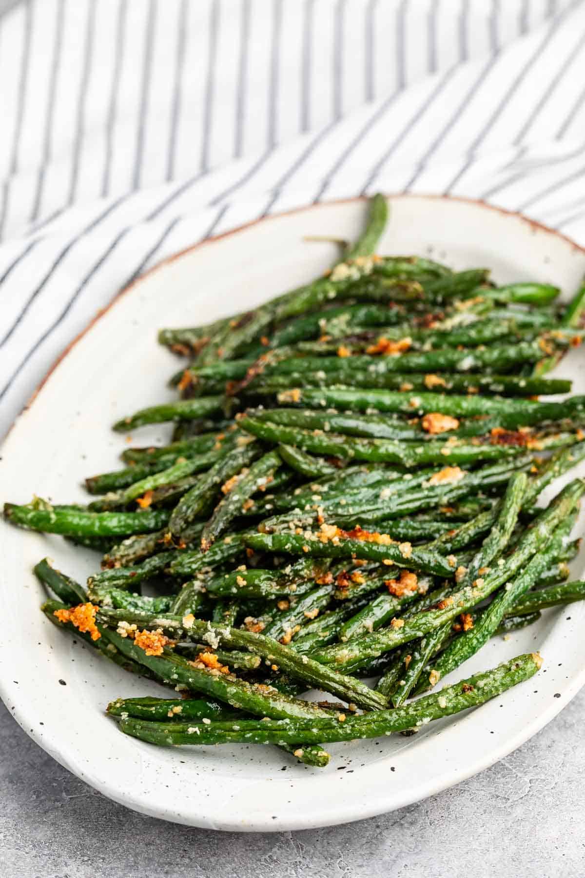 A white oval plate filled with roasted green beans topped with grated Parmesan cheese, set on a light surface with a striped cloth in the background.