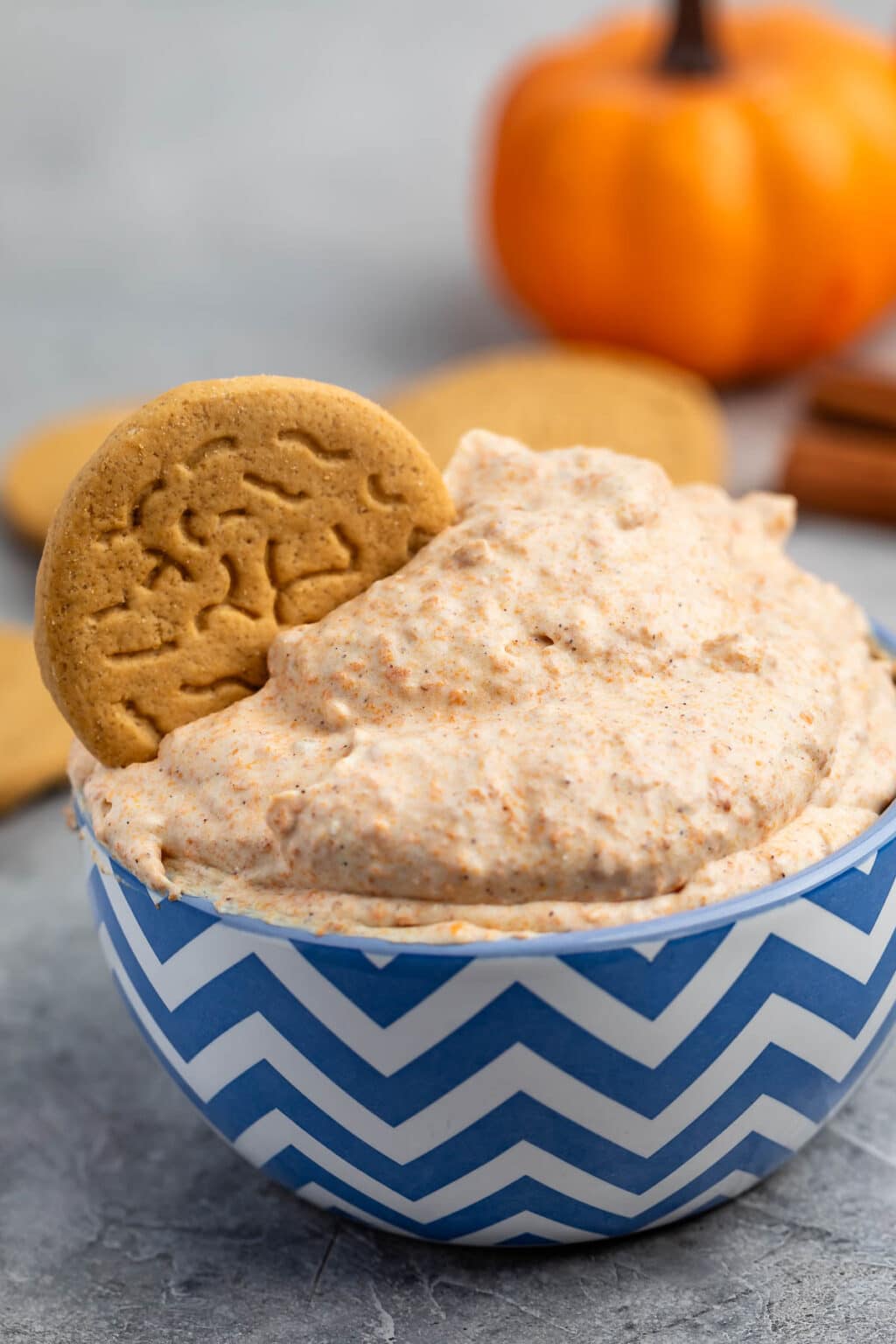 A blue and white chevron bowl filled with creamy pumpkin dip, with a round cookie dipped in it. A small pumpkin and cookies are blurred in the background.