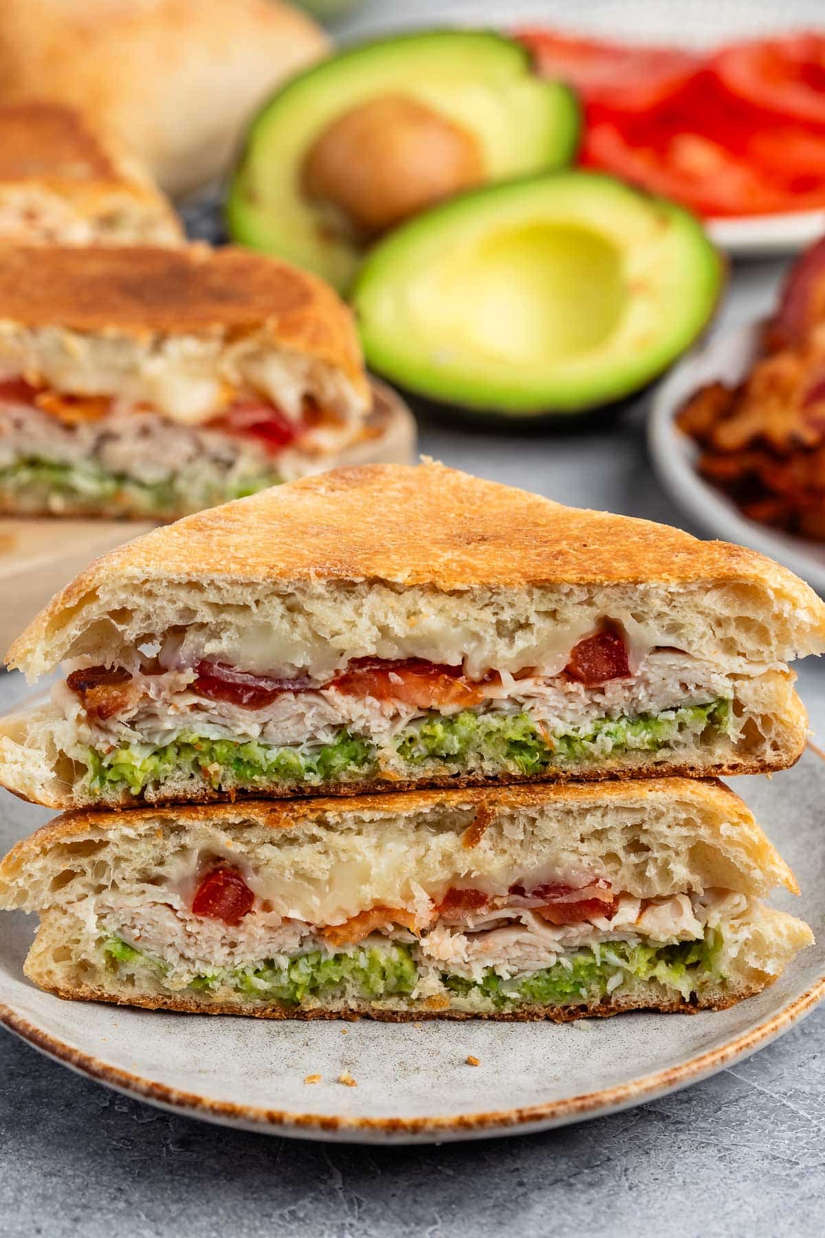 A toasted sandwich cut in half, showing layers of avocado, sliced turkey, melted cheese, tomato, and bacon on a plate, with avocado, tomatoes, and bacon in the background.