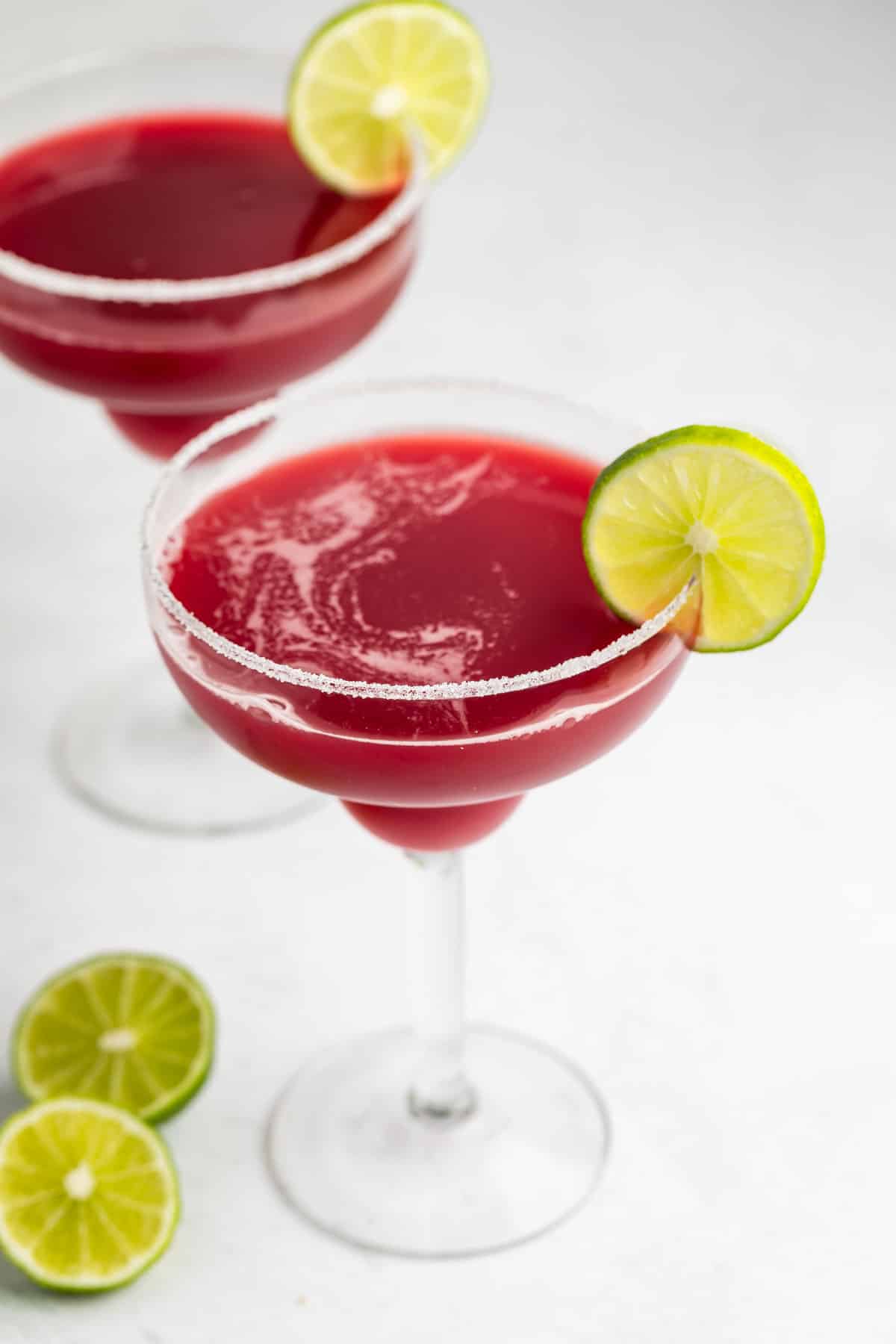 Two margarita glasses filled with a deep red cocktail, garnished with a lime wheel and a sugared rim. Slices of lime are scattered on the white surface around the glasses.