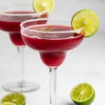 Two margarita glasses filled with a deep red drink, each garnished with a lime wheel on the rim. Lime halves are placed on the white surface nearby. The background is bright and minimal.
