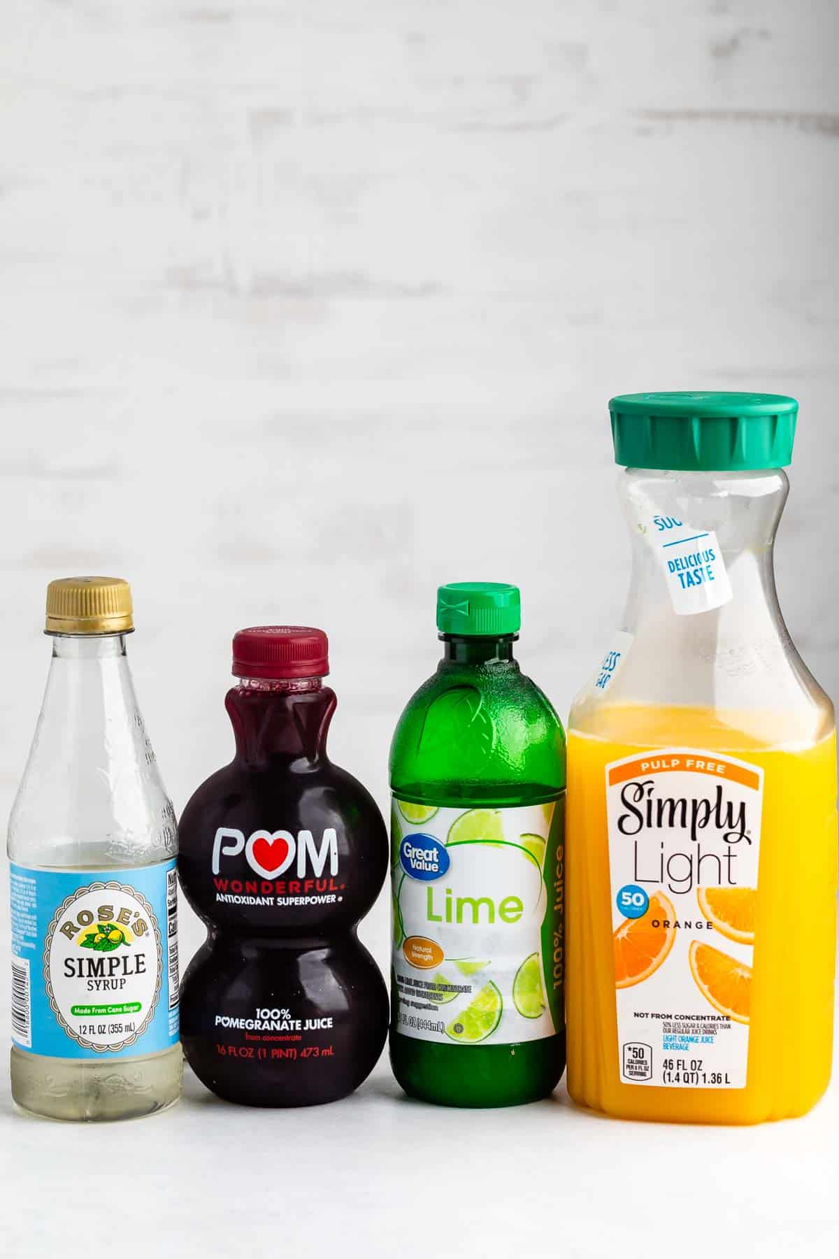 Four bottles stand side by side: simple syrup, pomegranate juice, lime juice, and a large Simply Light orange juice bottle, all against a white background.
