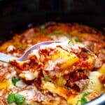 A spoon scoops cheesy, saucy lasagna from a slow cooker, showing layers of pasta, tomato sauce, melted cheese, and fresh herbs.