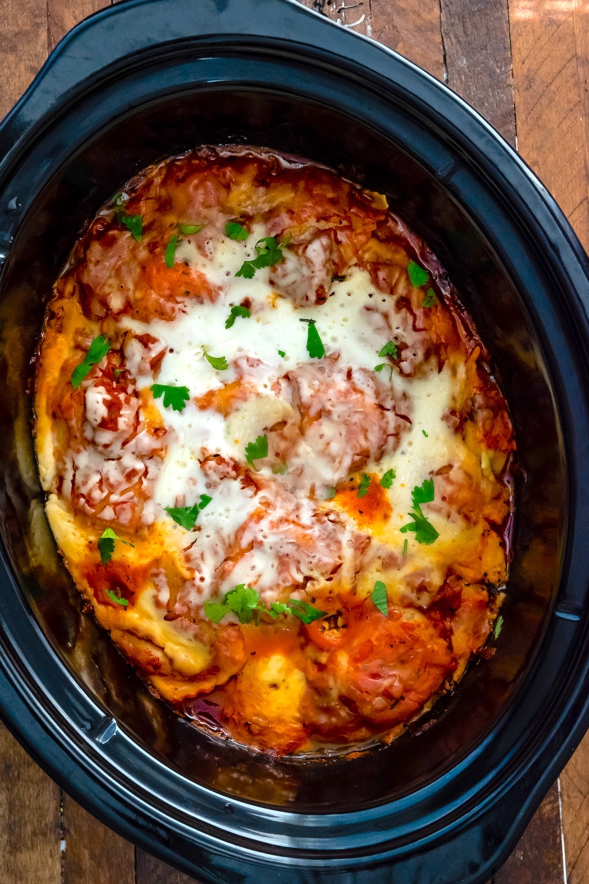 A close-up view of a homemade lasagna topped with melted cheese and fresh parsley, served in a black slow cooker on a wooden table.