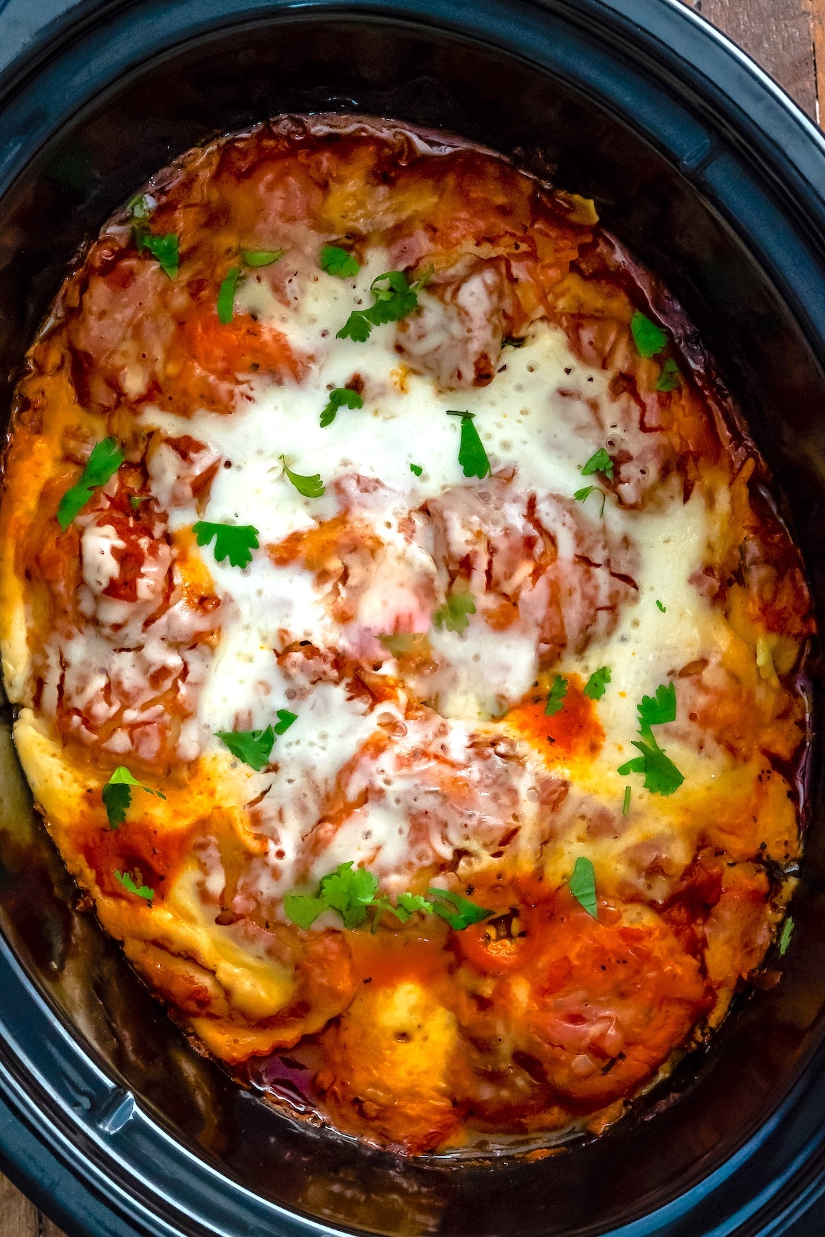 Lasagna topped with melted cheese and fresh parsley is cooked in a black slow cooker, with visible layers of sauce, pasta, and cheese.