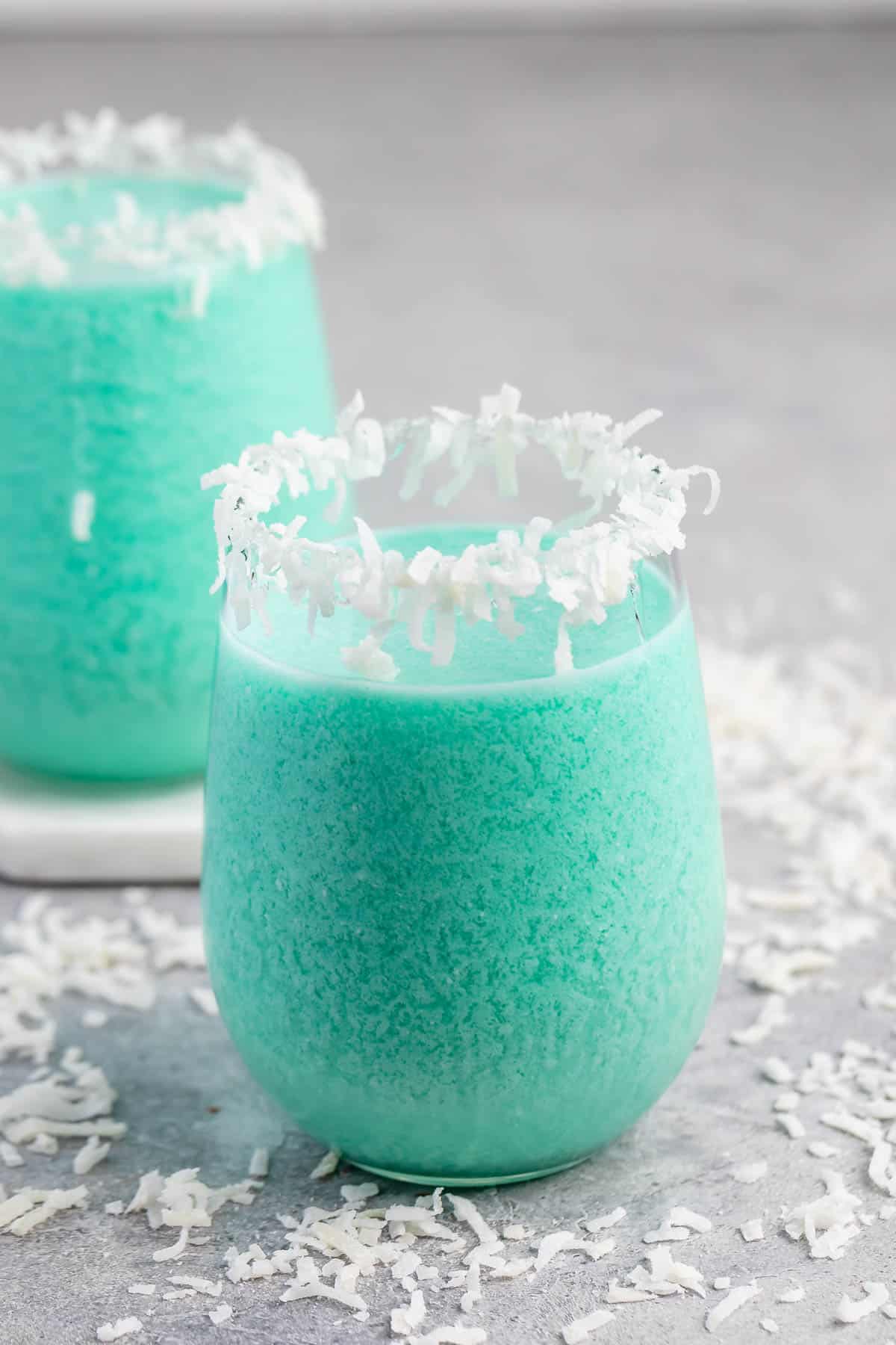 A glass of blue frozen drink with a rim coated in shredded coconut, surrounded by scattered coconut flakes on a gray surface. Another similar drink is blurred in the background.