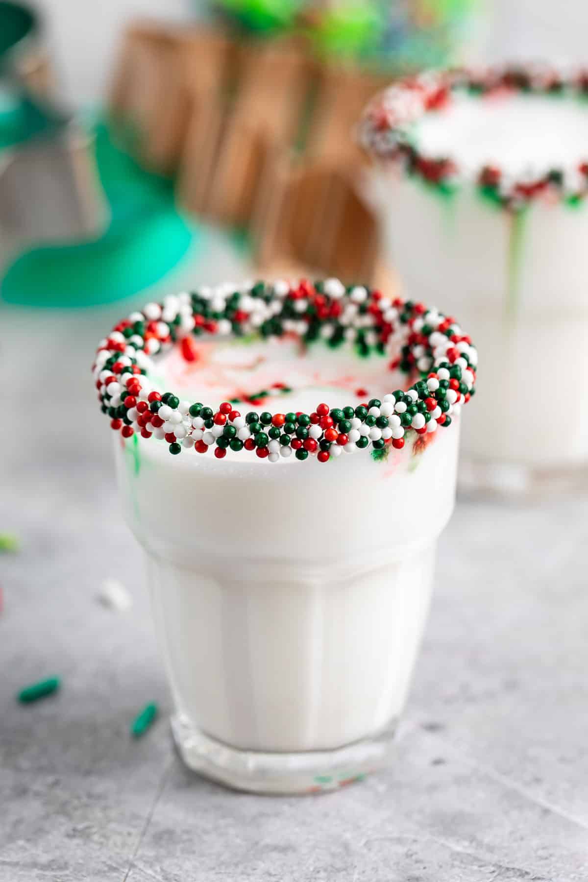 A glass of milk with the rim coated in red, green, and white sprinkles, sitting on a light gray surface. Another similar glass is blurred in the background.