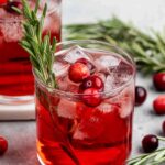 A glass of red cranberry cocktail with ice, garnished with fresh cranberries and a sprig of rosemary, sits on a gray surface with scattered cranberries and rosemary sprigs nearby.