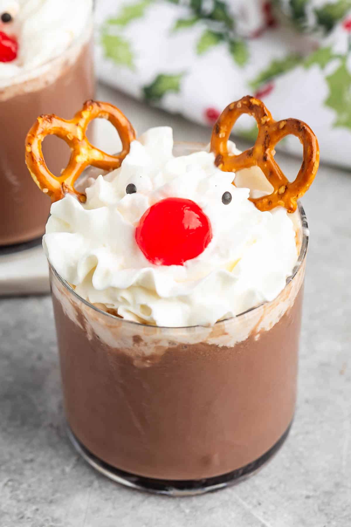 A glass of hot chocolate topped with whipped cream, a red cherry for a nose, two small pretzels as antlers, and two chocolate dots for eyes, decorated to look like a reindeer.