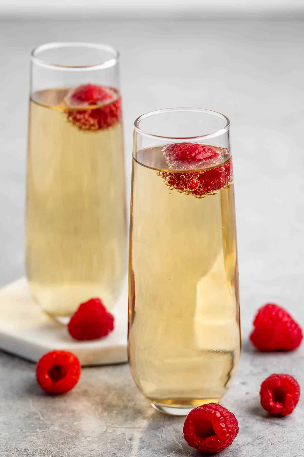 Two champagne flutes filled with sparkling wine, each garnished with a fresh raspberry. Several loose raspberries are placed on the gray surface around the glasses.