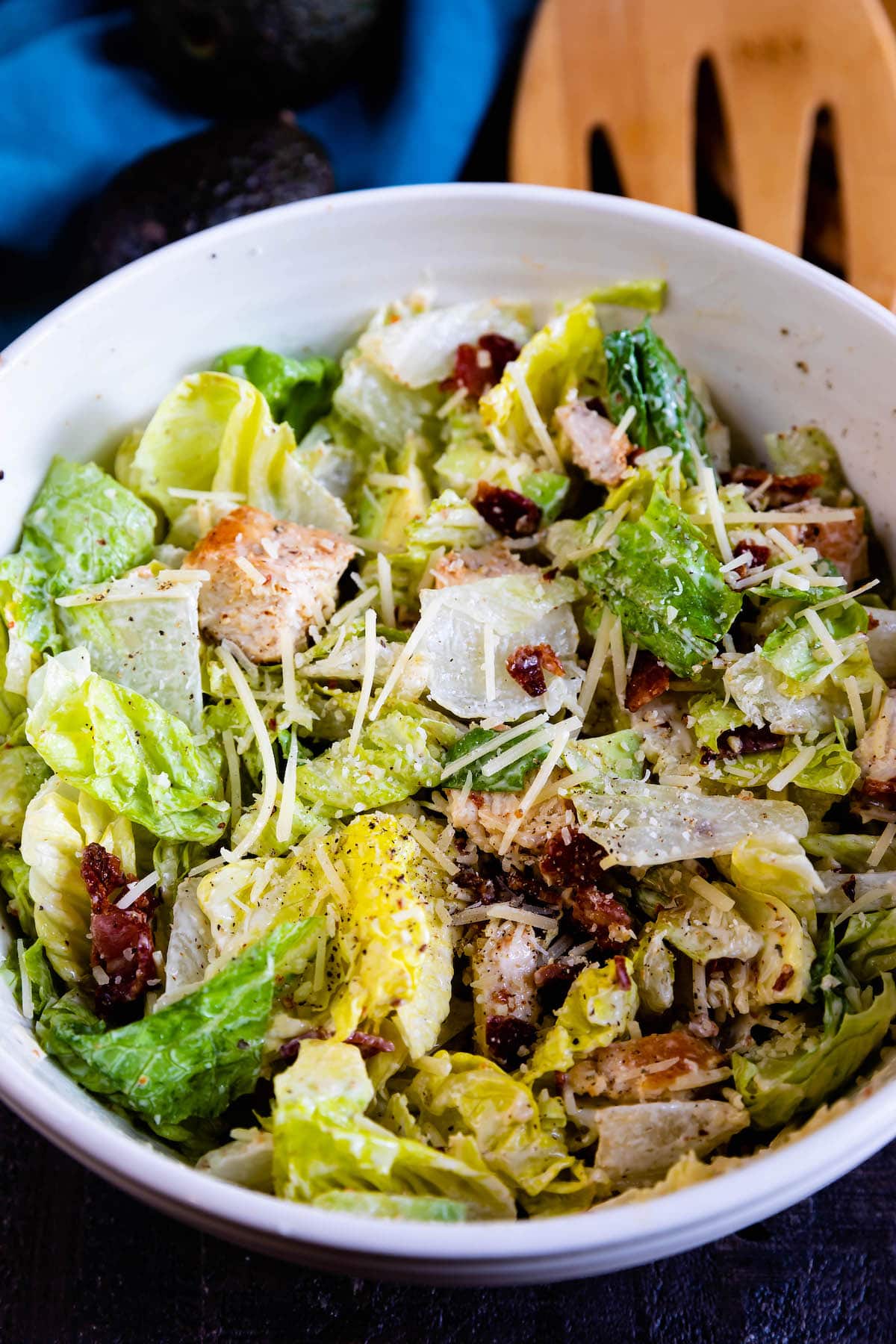 A bowl of Caesar salad with chopped romaine lettuce, grilled chicken pieces, shredded Parmesan cheese, and bits of bacon, all tossed in creamy dressing. A wooden salad server is partially visible in the background.