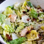 A bowl of Caesar salad with romaine lettuce, grilled chicken pieces, grated Parmesan cheese, and crispy bacon bits, garnished with cracked pepper. Wooden salad tongs are visible in the background.