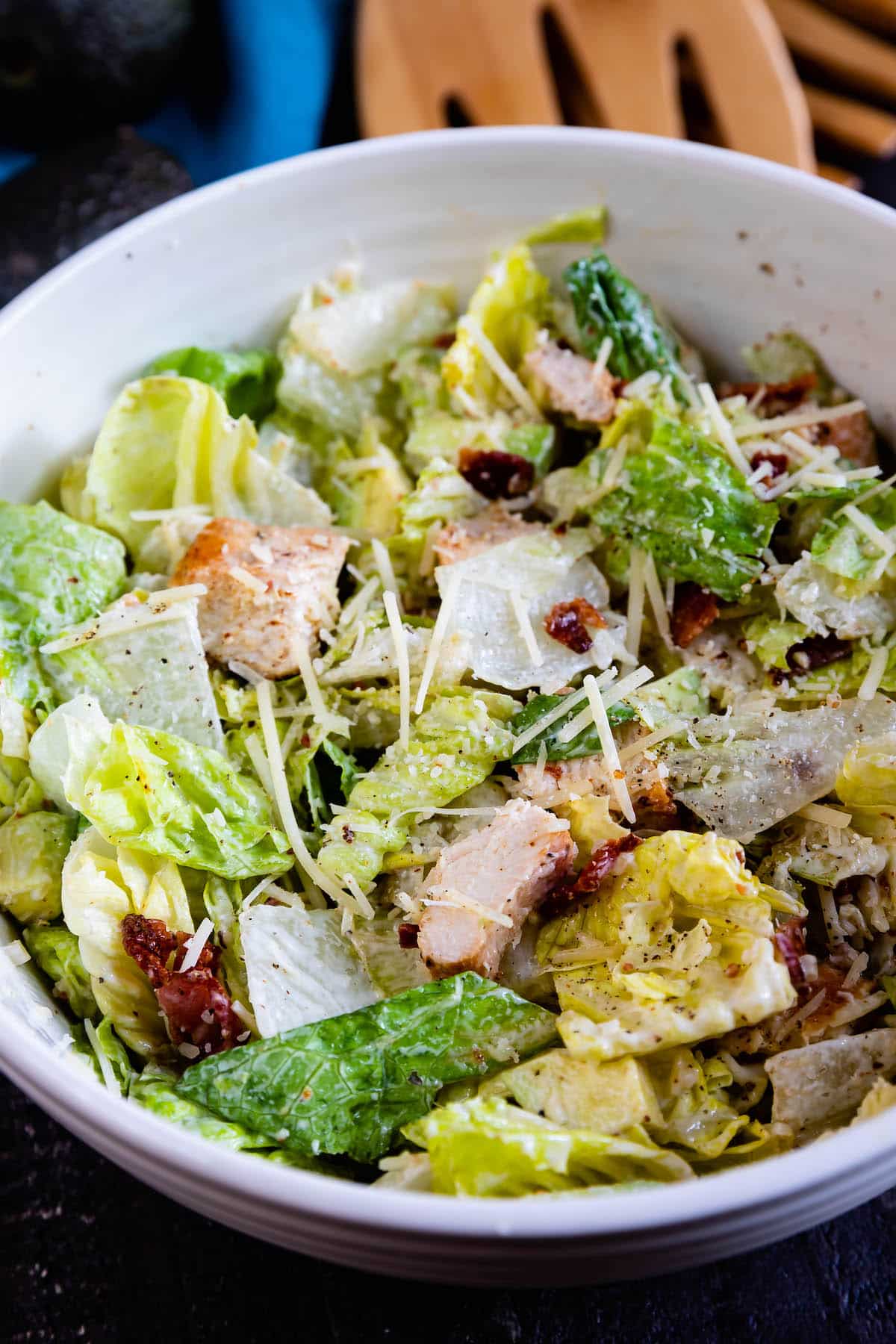 A bowl of Caesar salad with romaine lettuce, grilled chicken pieces, grated Parmesan cheese, and crispy bacon bits, garnished with cracked pepper. Wooden salad tongs are visible in the background.