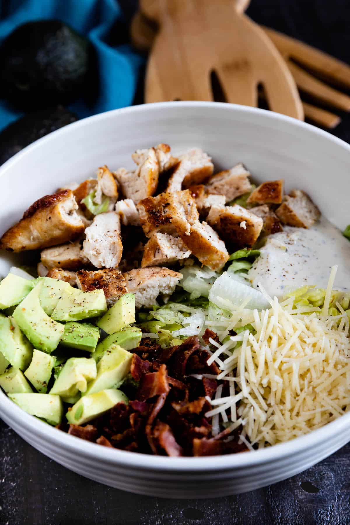 A white bowl filled with chopped grilled chicken, diced avocado, bacon bits, shredded cheese, lettuce, and a dollop of creamy dressing, with wooden salad servers and avocados in the background.
